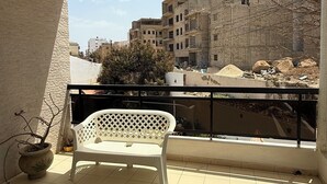 Un balcone con una sedia di plastica bianca, un letto fatto con un pneumatico giallo e vista sugli edifici sullo sfondo.