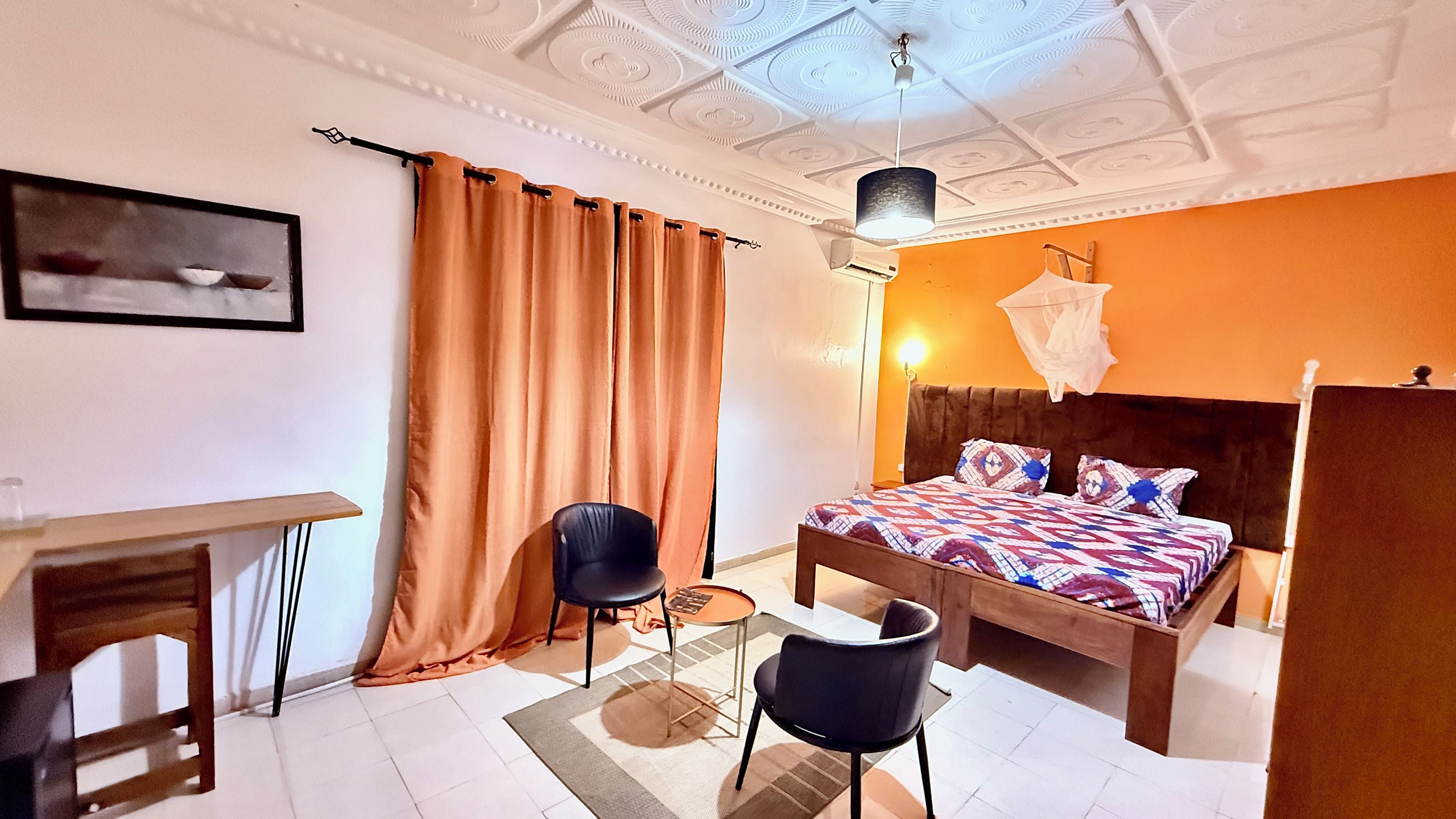 A bedroom with a bed, a desk, two chairs, a ceiling fan, and a window with curtains.