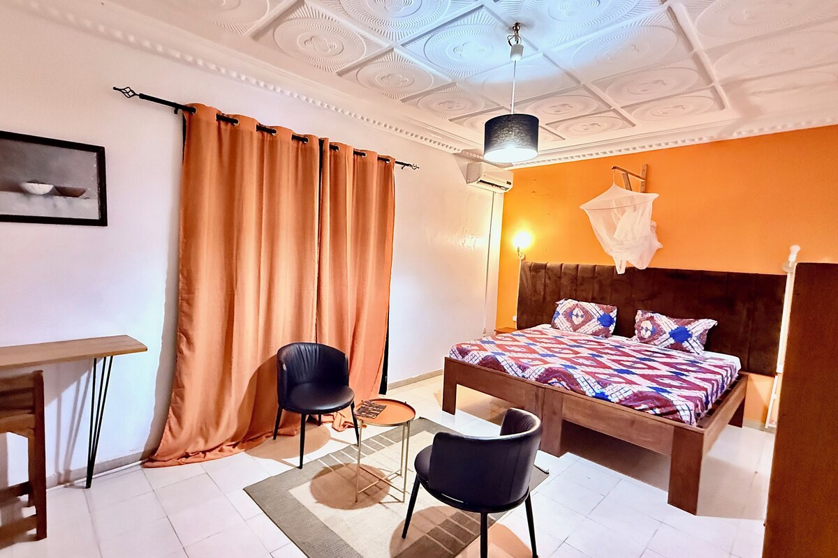 A bedroom with a bed, a desk, two chairs, a ceiling fan, and a window with curtains.