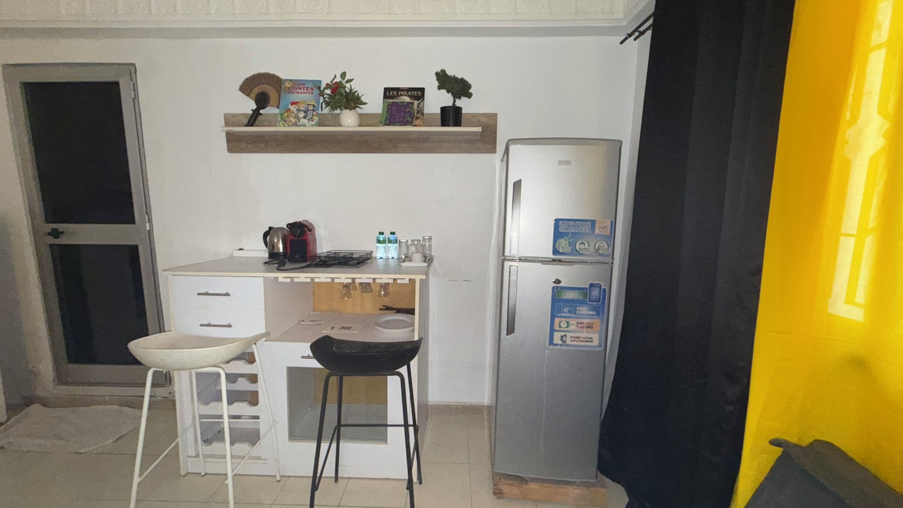 Una kitchenette compacta con refrigerador, una pequeña encimera, dos taburetes, un lavamanos y un estante con libros y una planta.