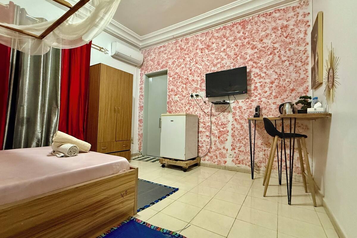 A bedroom with a bed, a wardrobe, a desk with a chair, a television, and a refrigerator.