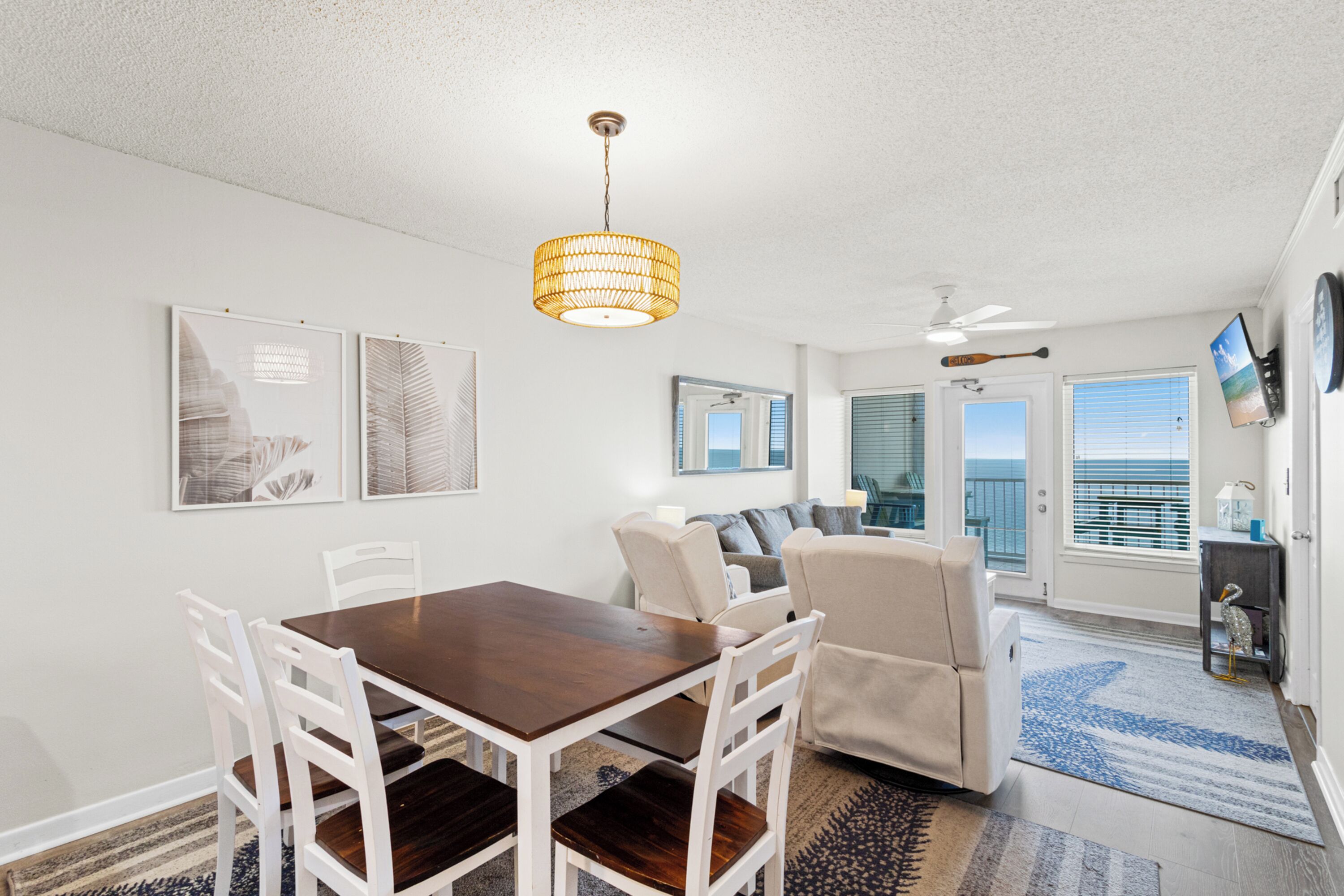 A dining area with a wooden table and white chairs, a ceiling fan, and a view of the ocean through the sliding glass doors.