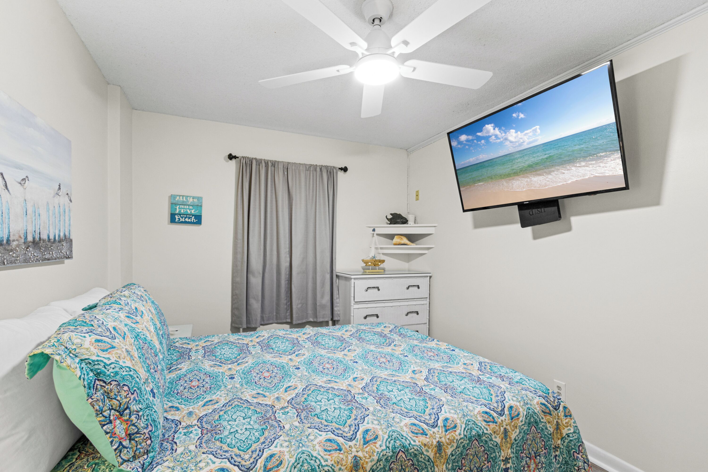 A bedroom with a bed, a ceiling fan, a television displaying a beach scene, a nightstand, and a wall with a painting and a sign.