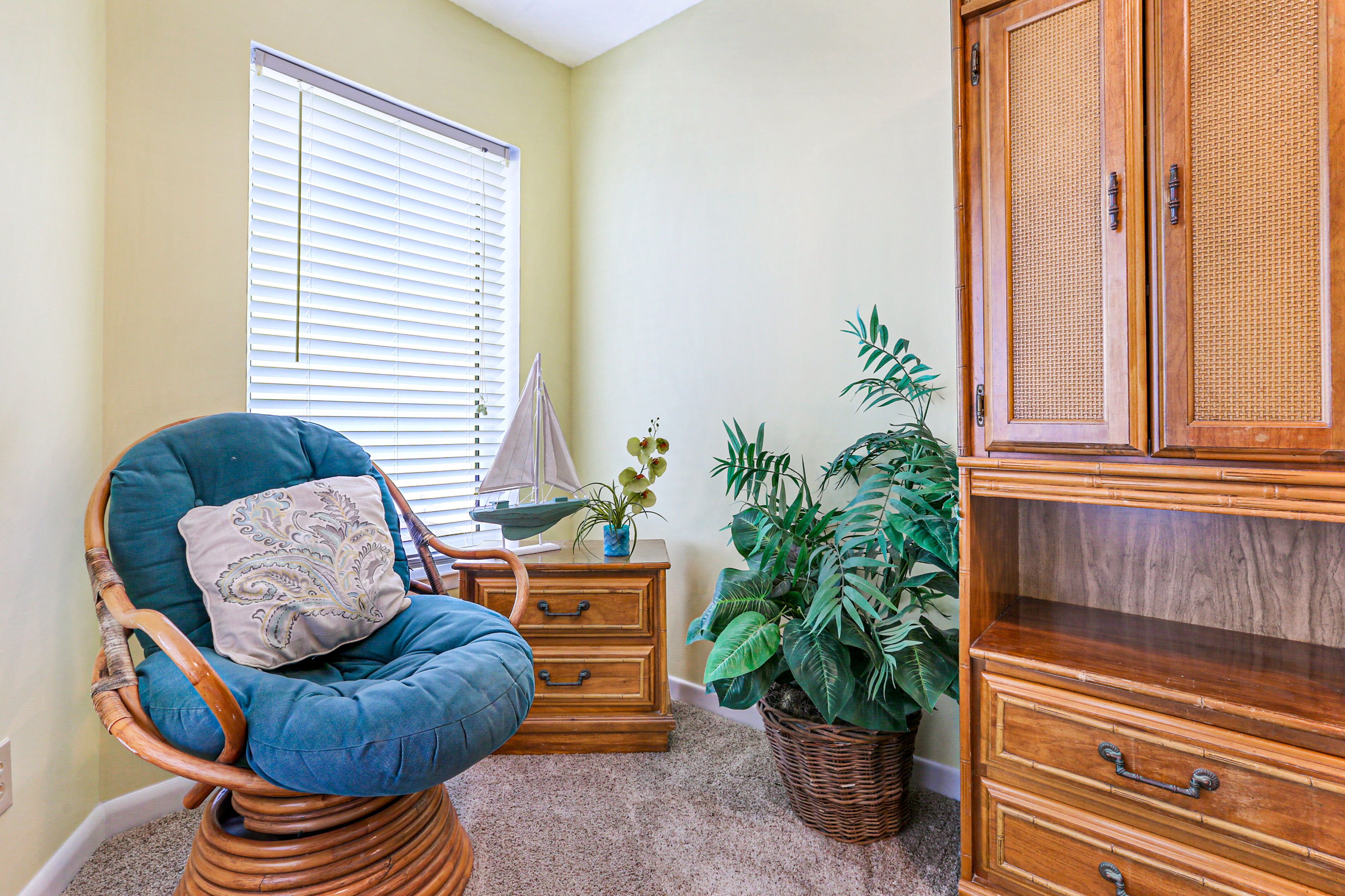 A room with a wicker chair, a wooden cabinet, a potted plant, and a window with blinds.
