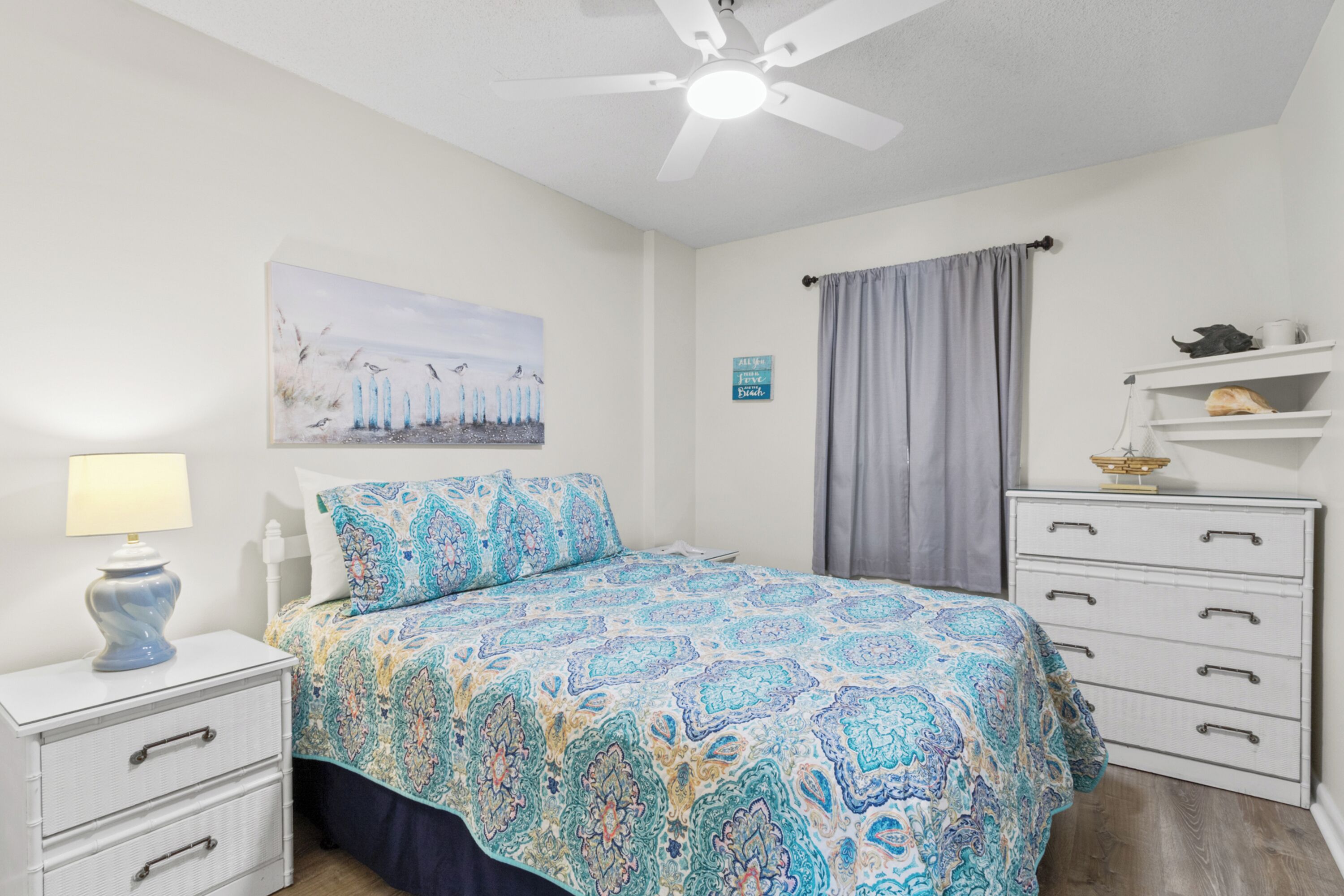 A bedroom with a bed, a nightstand, a lamp, a ceiling fan, a painting, a shelf, and a curtain.