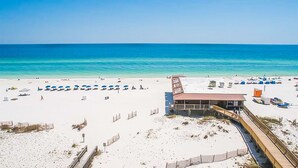 A beach with a boardwalk leading to a building with a red roof, white walls, and a sign that reads "The Beach House." - Boardwalk Condos by Hosteeva (Gulf Shores)
