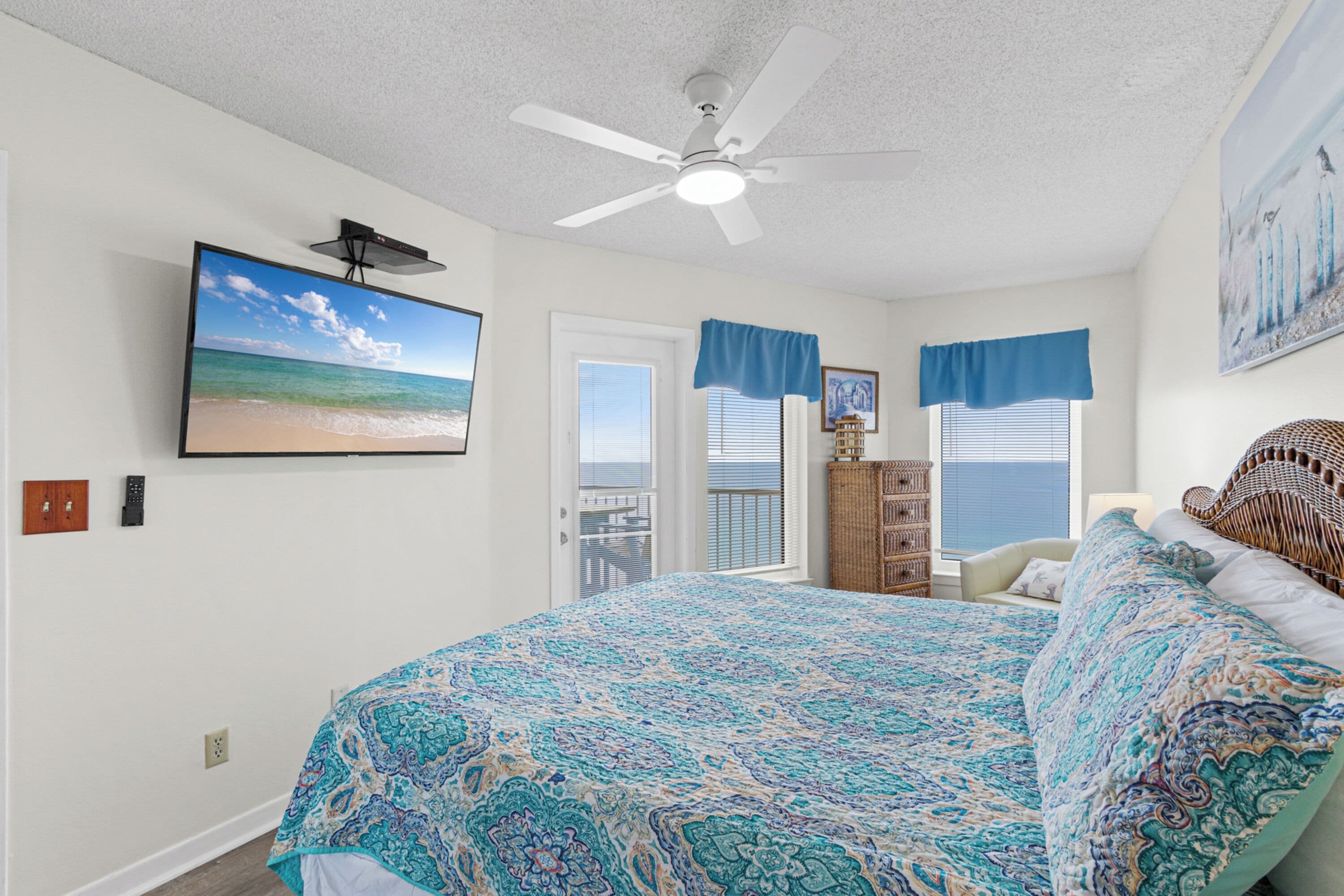 A bedroom with a large bed, a ceiling fan, a TV mounted on the wall, and a view of the ocean through the window.