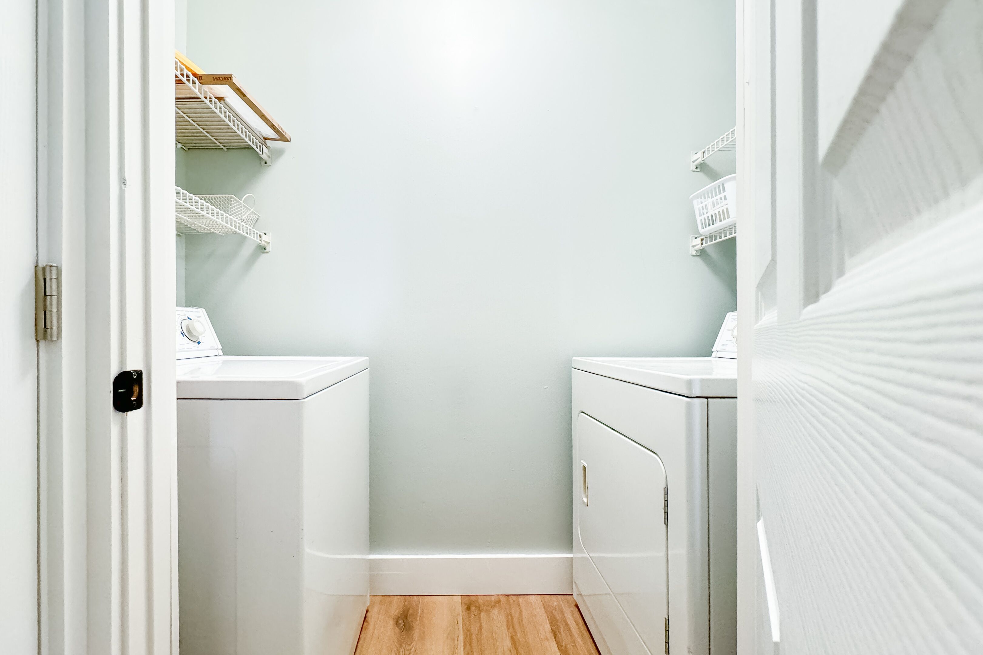 A laundry room with two white washing machines, wooden flooring, and white shelving units with baskets.