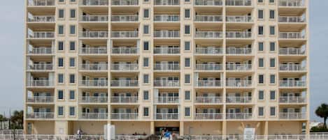Un hôtel à plusieurs étages, à l’architecture extérieure uniforme, doté de balcons à chaque étage et bordé d’une plage de sable.