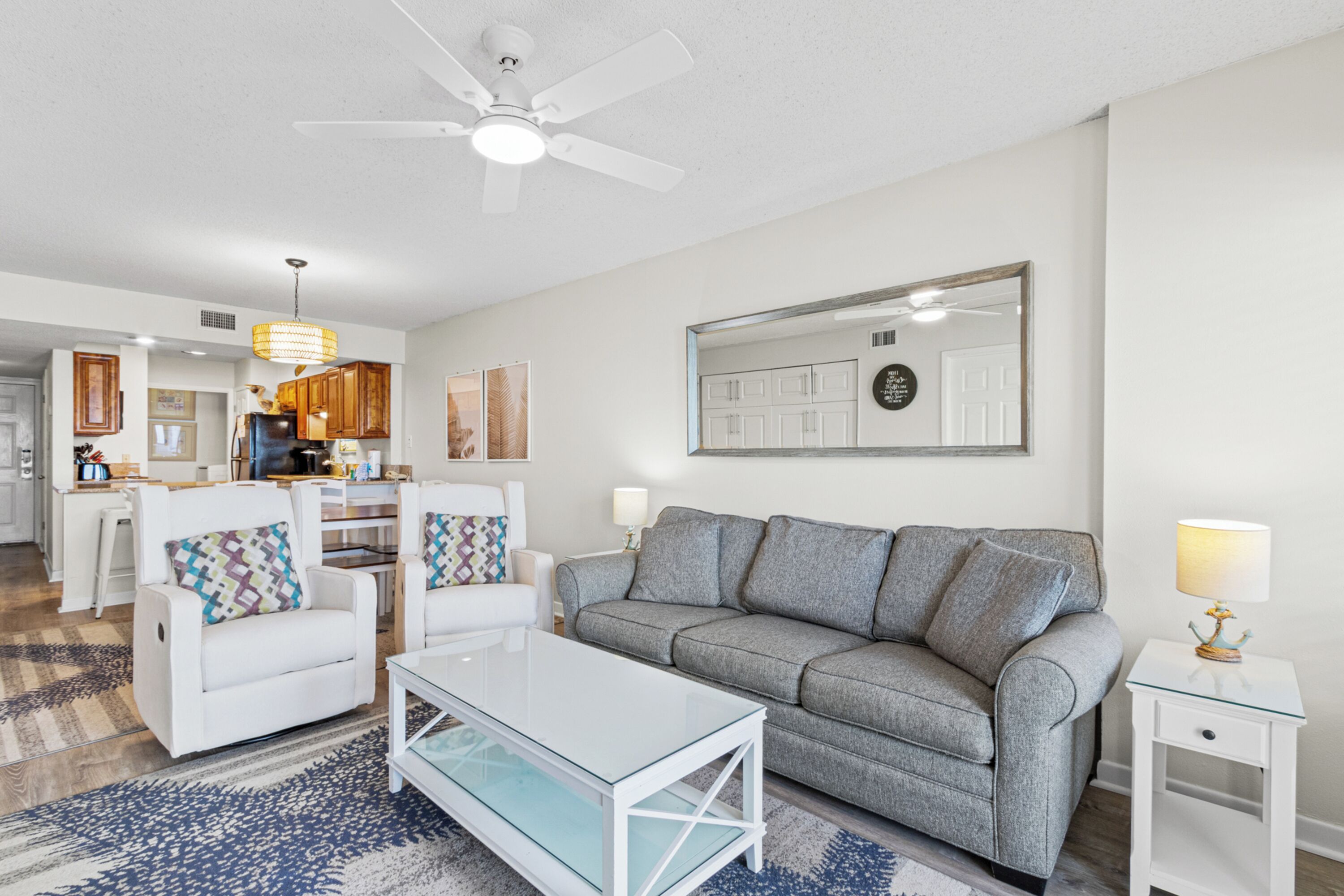 A modern living room with a grey sofa, white armchairs, a glass coffee table, and a ceiling fan.