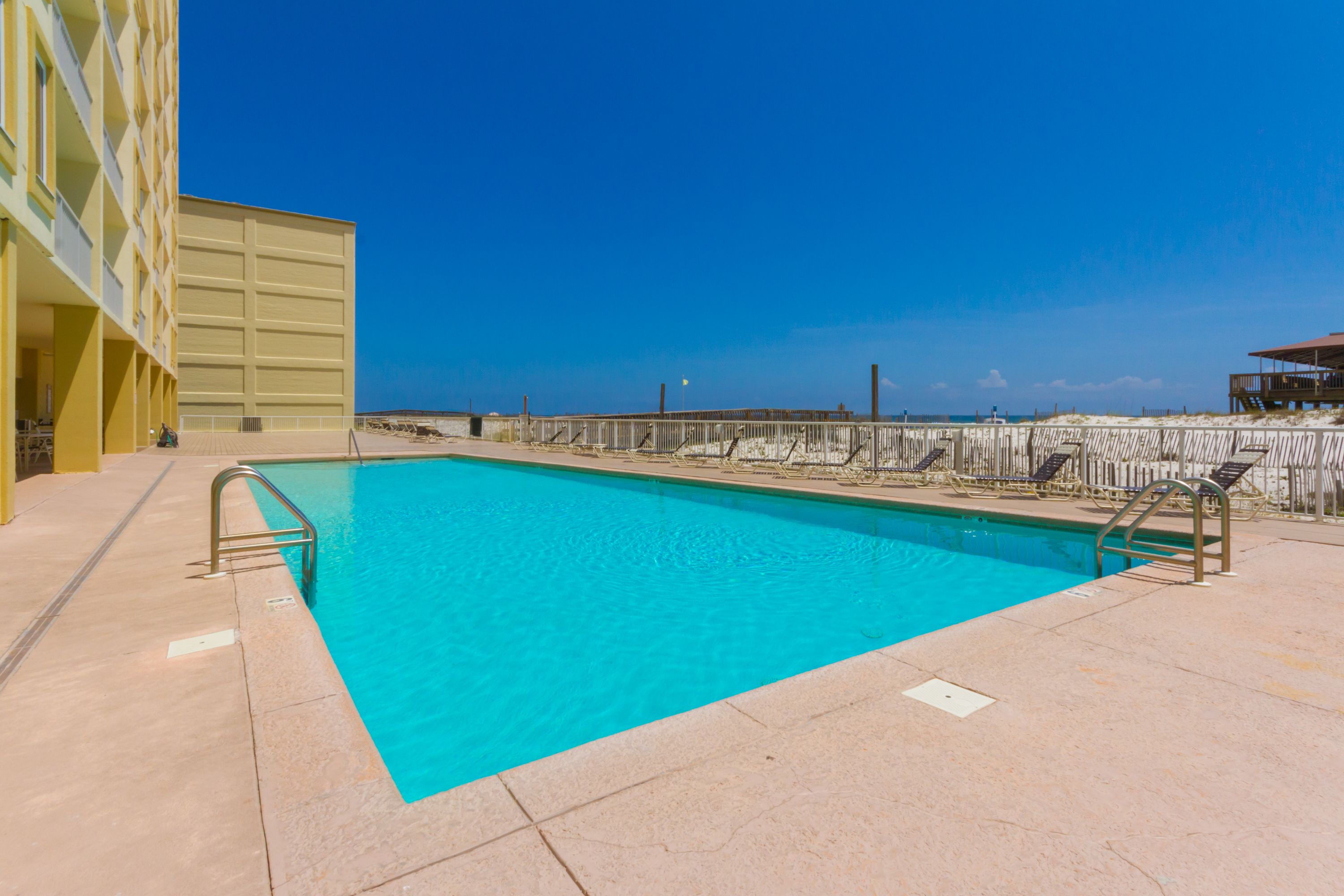 Une piscine à l’eau d’un bleu limpide, entourée d’une terrasse en bois et bordée d’un bâtiment doté de balcons.