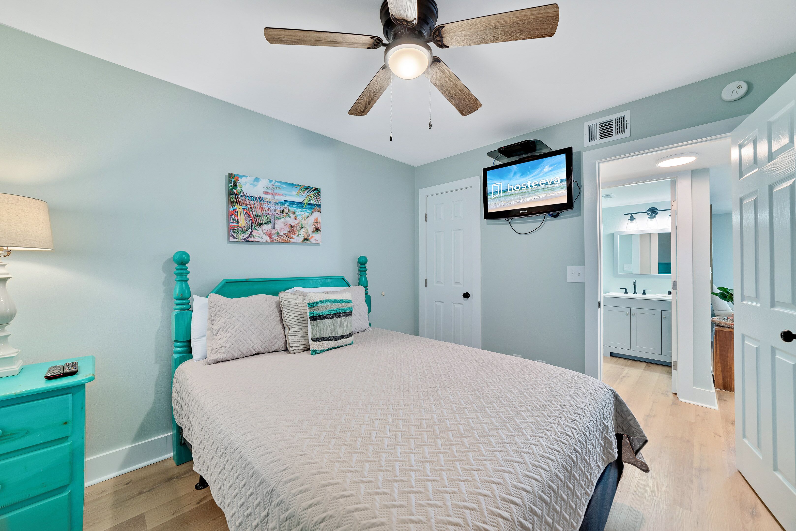 A bedroom with a bed, a ceiling fan, a TV, and a door leading to a bathroom.