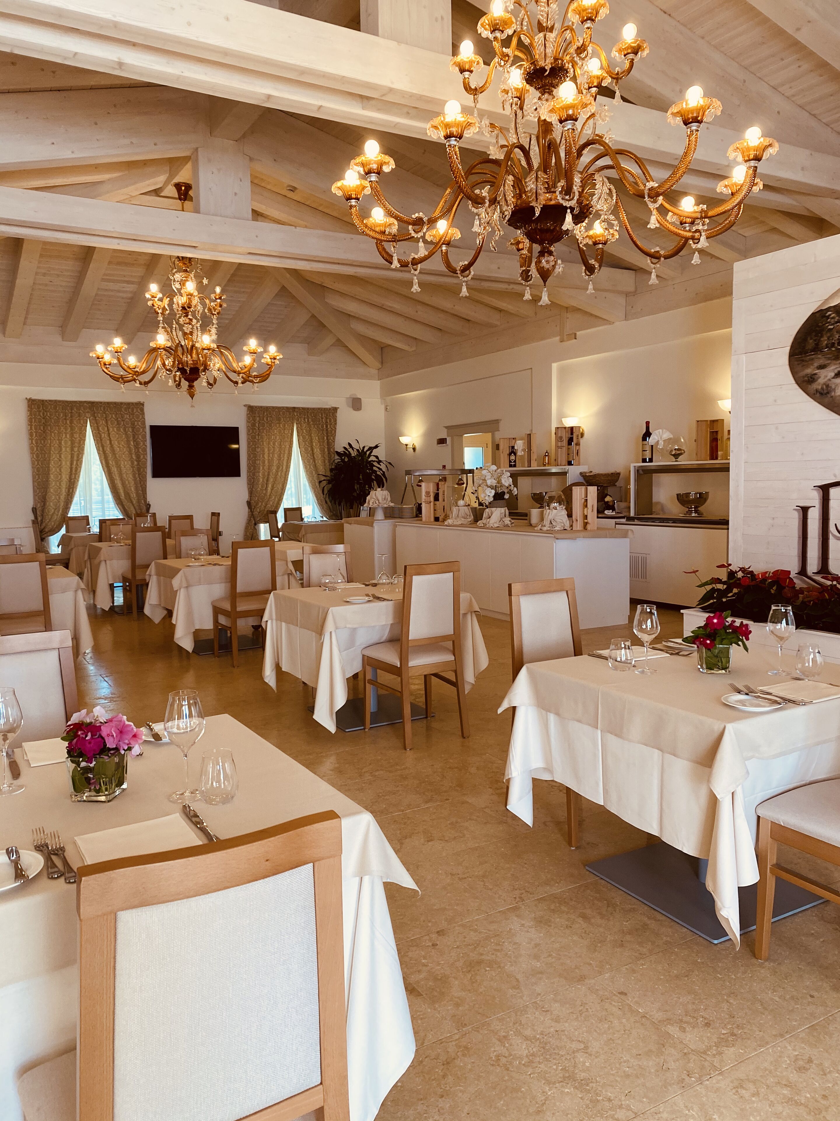 Een restaurant met meerdere eettafels gedekt voor gasten, een kroonluchter en een keuken in de achtergrond.