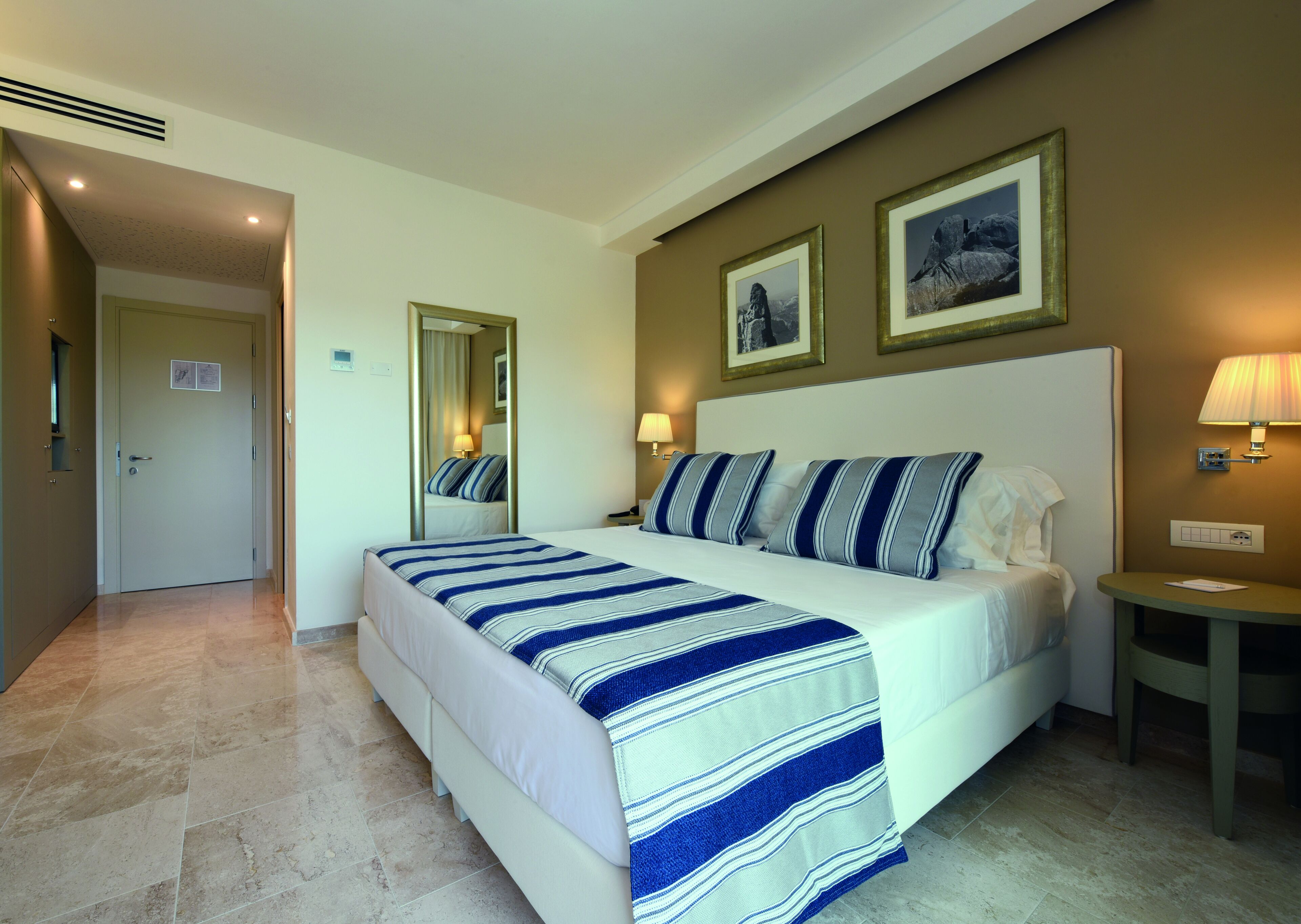 Habitación de hotel con una cama grande, una mesita de noche con lámpara, un espejo y dos cuadros enmarcados en la pared.