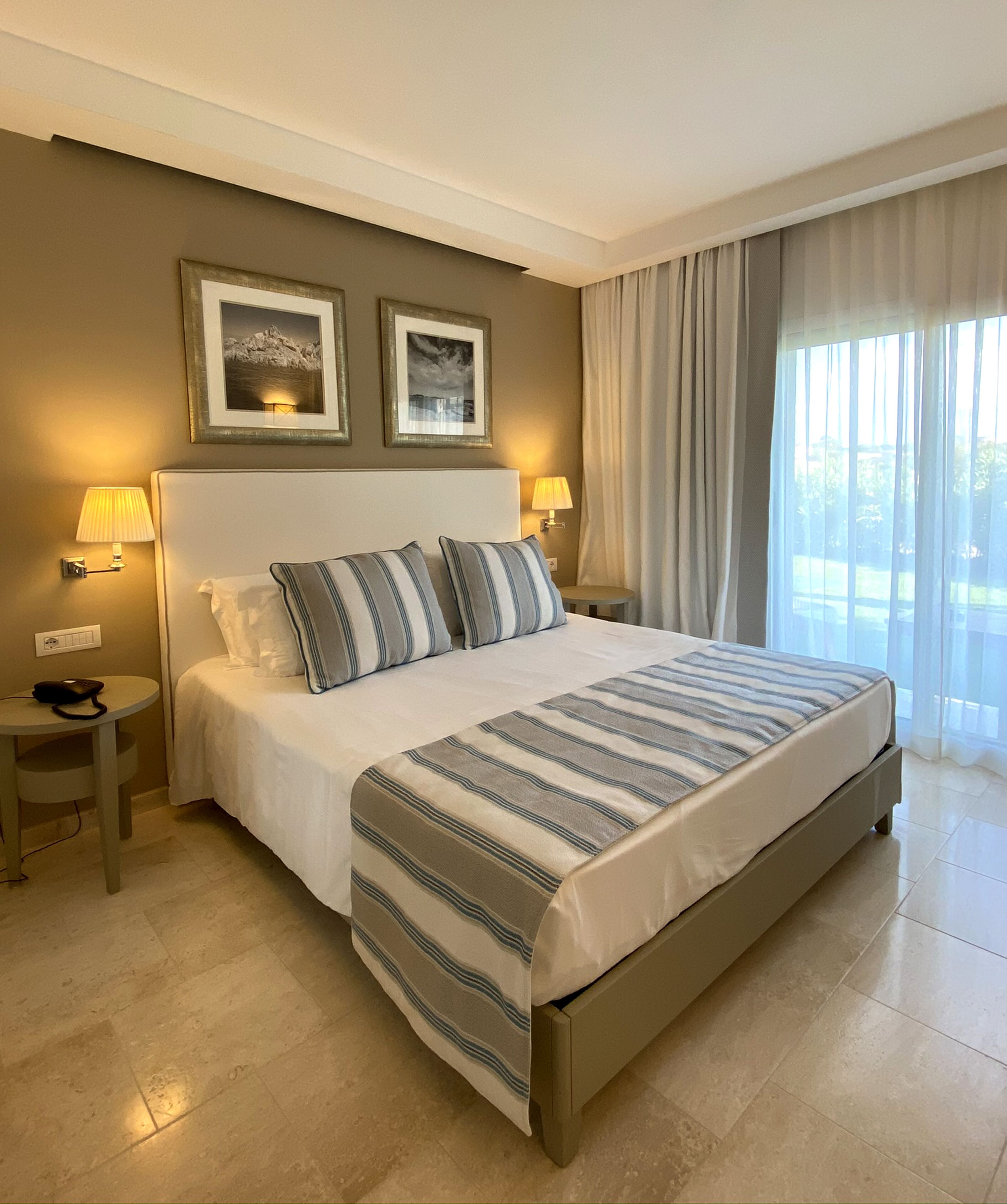 A hotel room with a large bed, two bedside lamps, a small table, and a window with curtains.