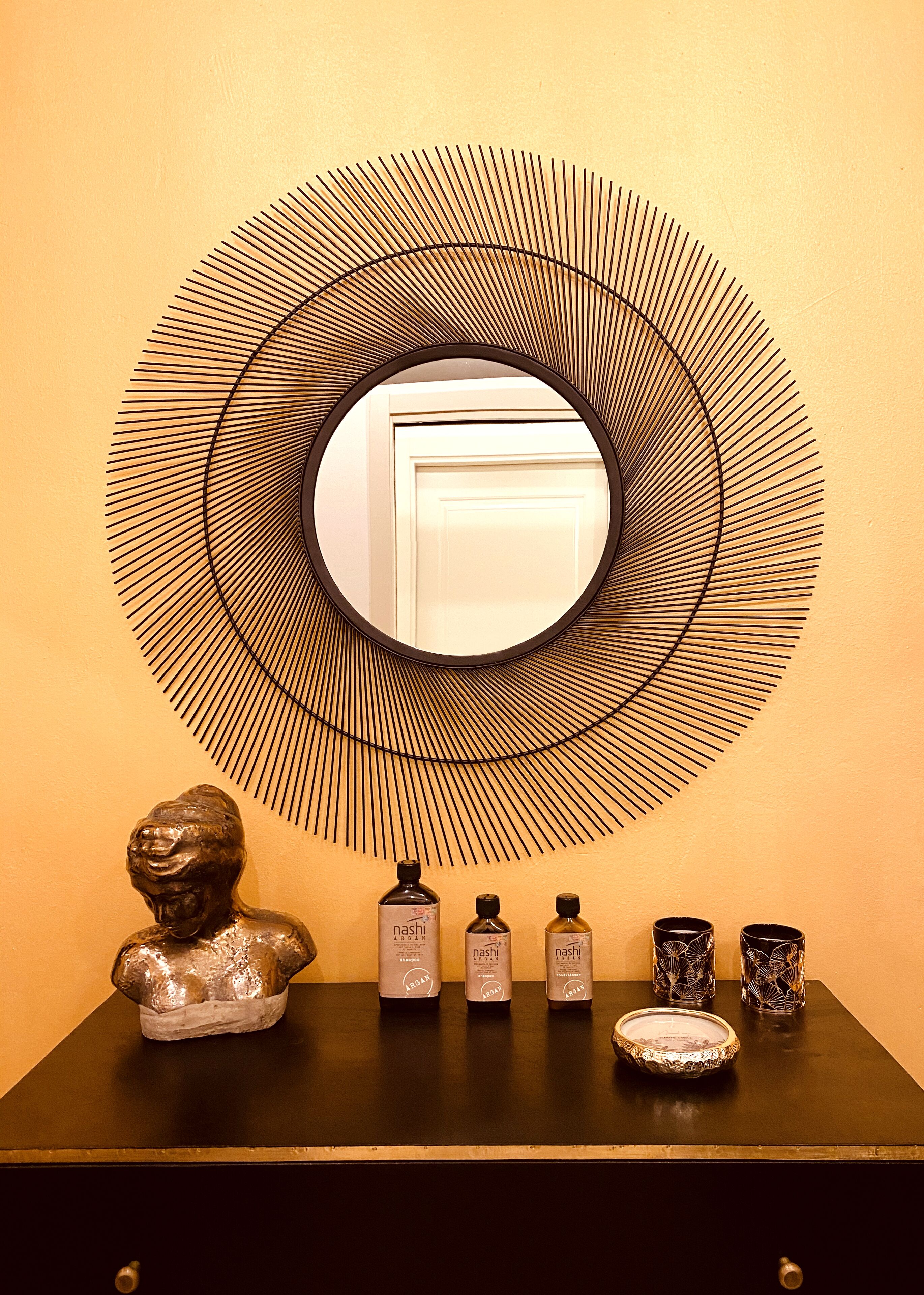 A wall-mounted mirror with concentric circles, a bust sculpture, and three bottles labeled 'nashi' on a black surface.