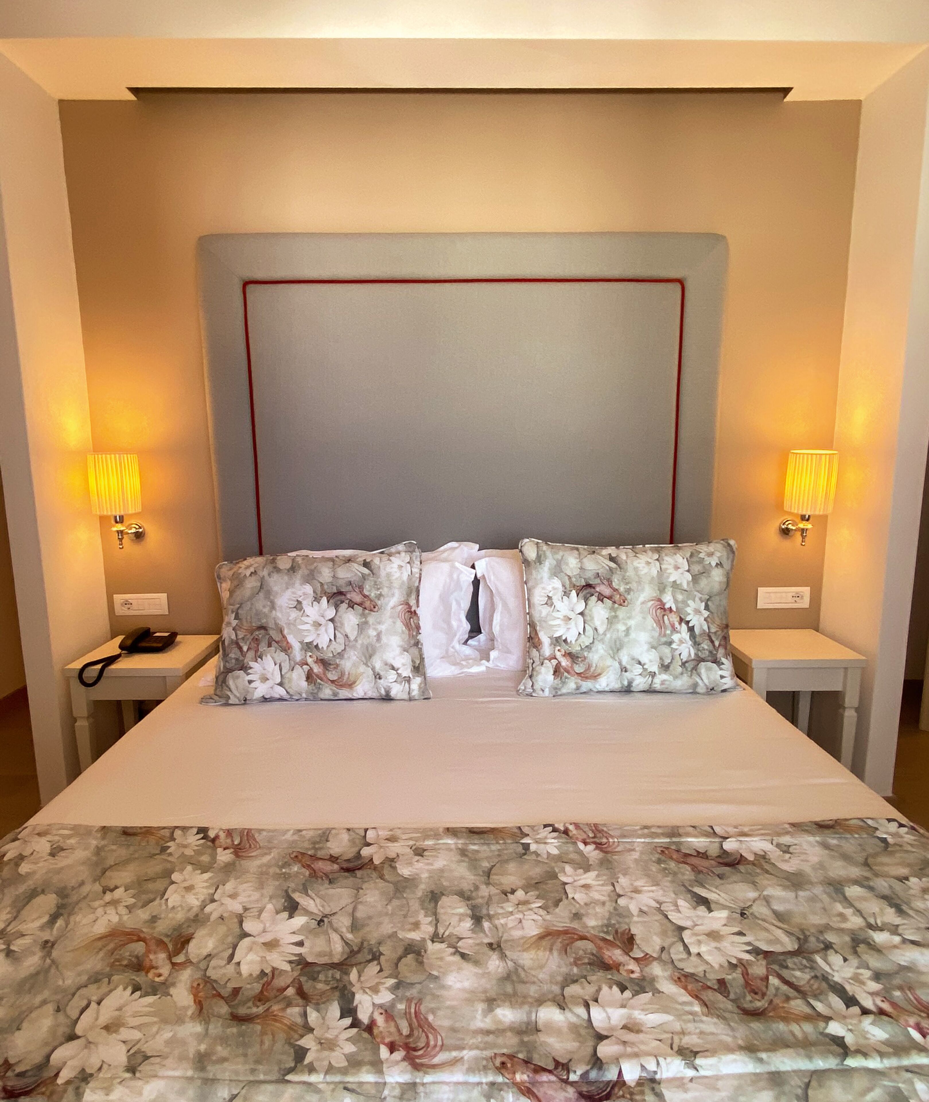 A hotel room with a large bed, floral-patterned pillows, two bedside tables with lamps, and a plain headboard.