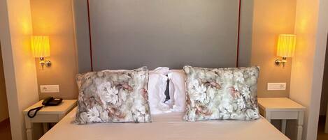 A hotel room with a large bed, floral-patterned pillows, two bedside tables with lamps, and a plain headboard.