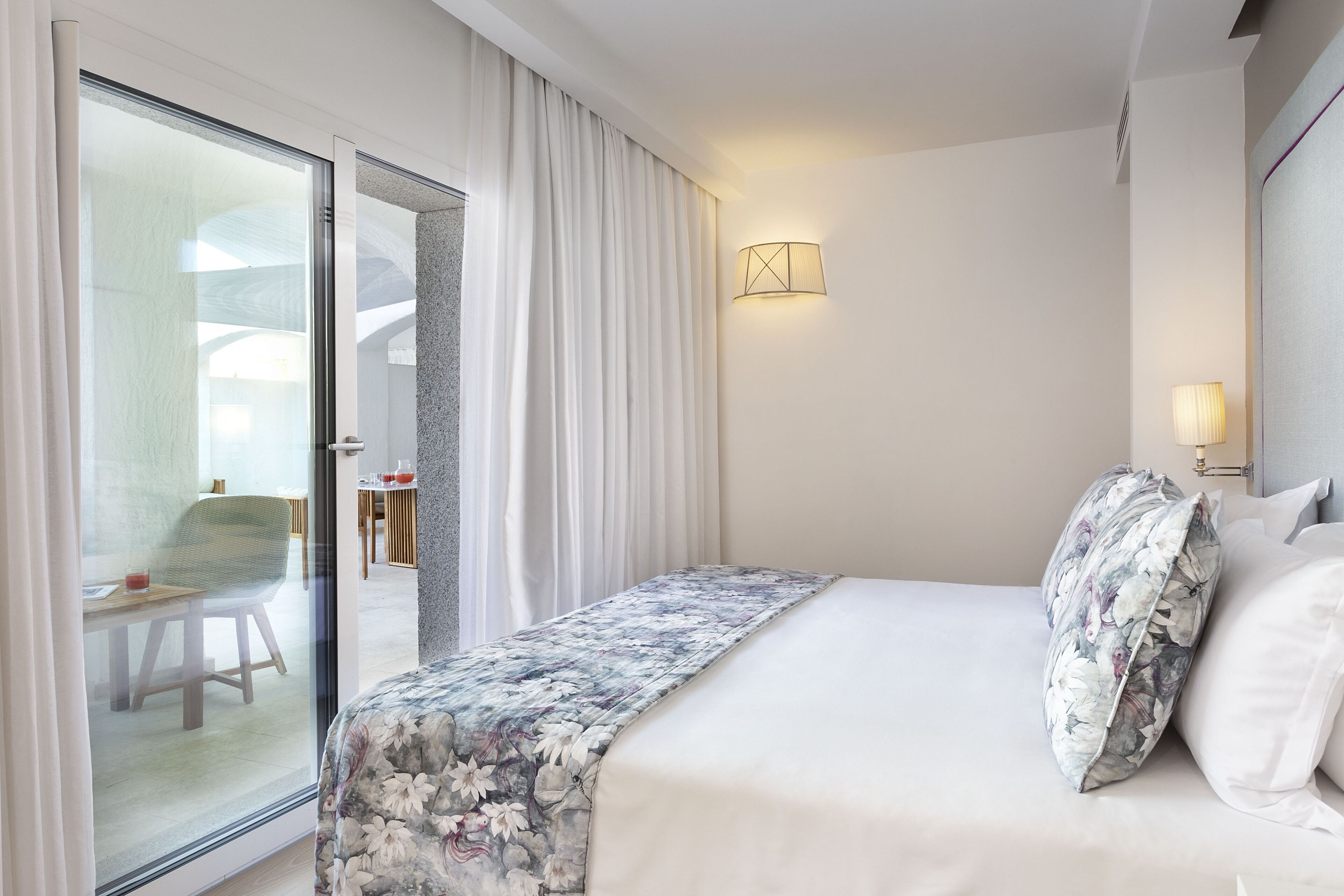 Una camera d'albergo con un letto, un comodino, una lampada a parete e una porta scorrevole in vetro che dà su un'area pranzo.