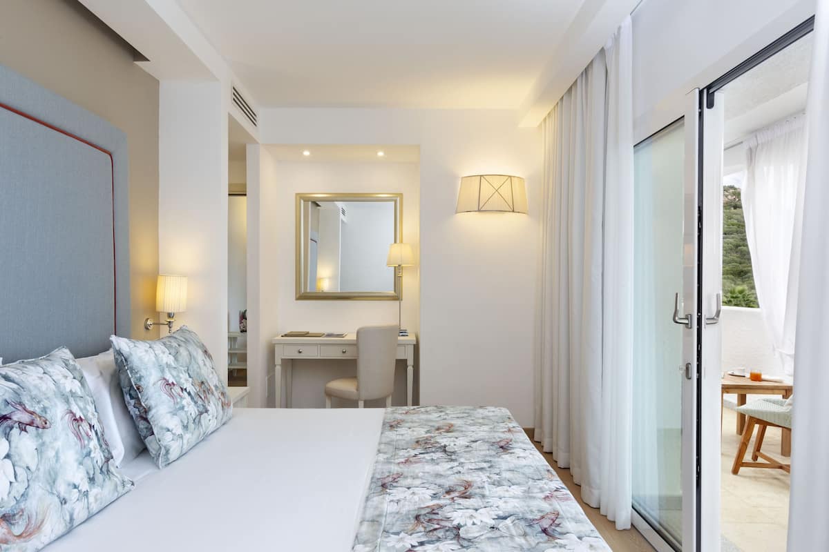 A hotel room with a bed, a desk, a chair, a mirror, and a view through a glass door.