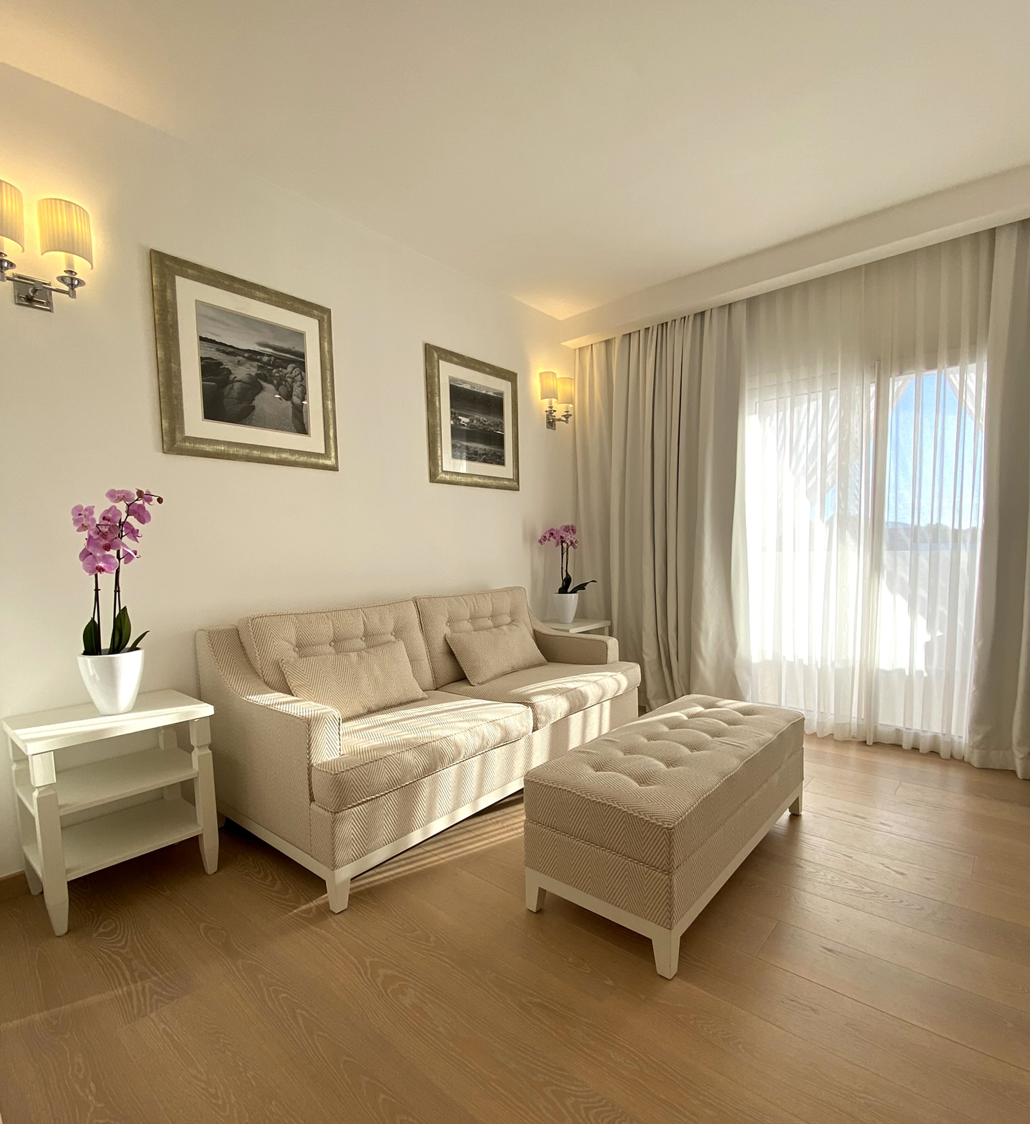 A modern living room with a beige sofa, a matching ottoman, and two nightstands with lamps. There are two framed pictures on the wall and a large window with sheer curtains.