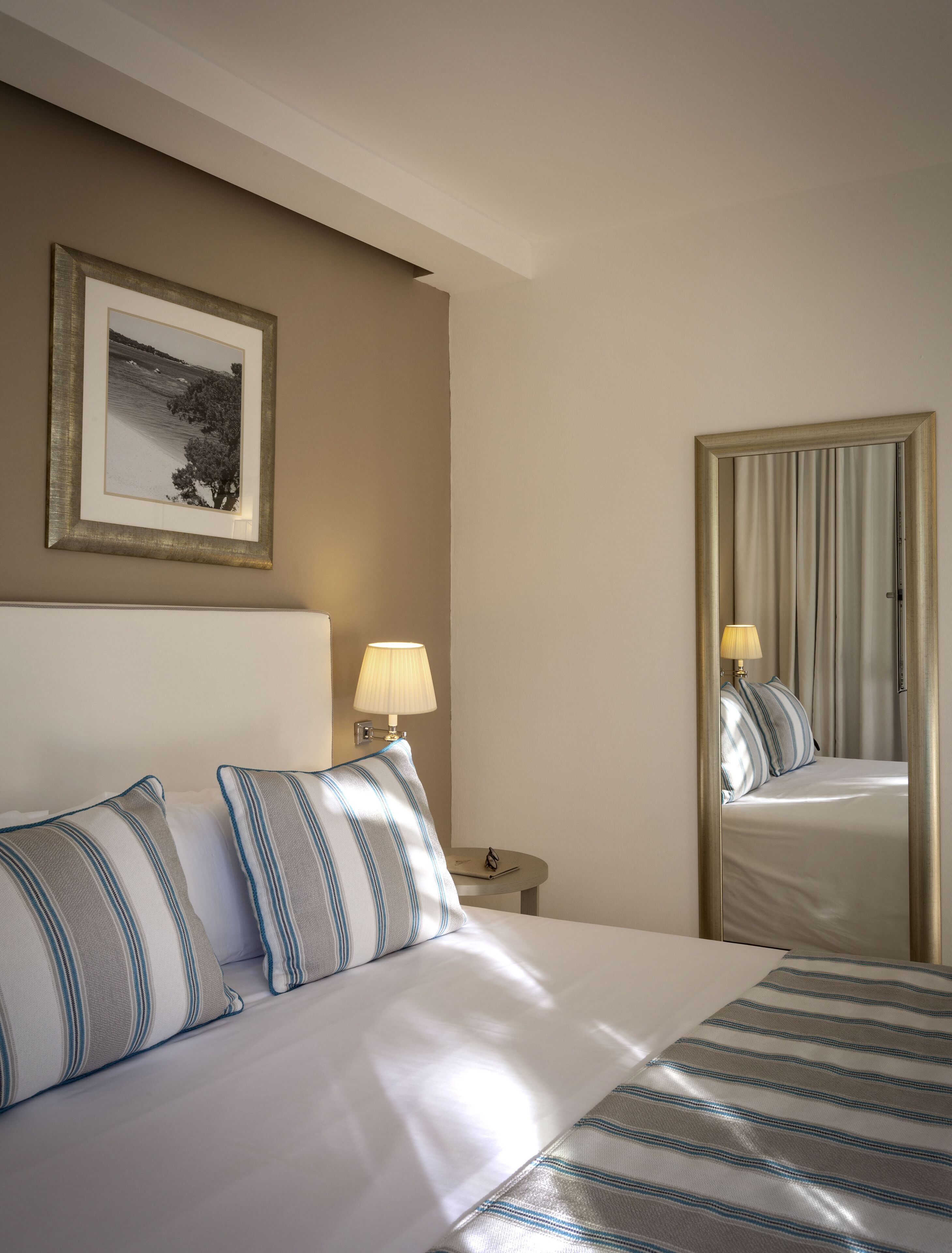 A hotel room with a bed, a nightstand, a lamp, a framed picture on the wall, and a mirror reflecting the bed and lamp.