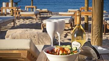 A wooden table with a bowl of salad, a bottle of wine, and a white wine glass on a beach setting.