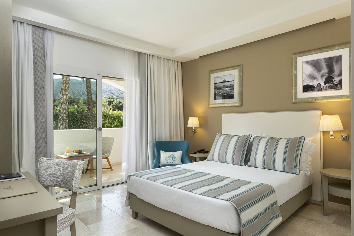 A hotel room with a large bed, a desk, a chair, and a balcony with a view of greenery.
