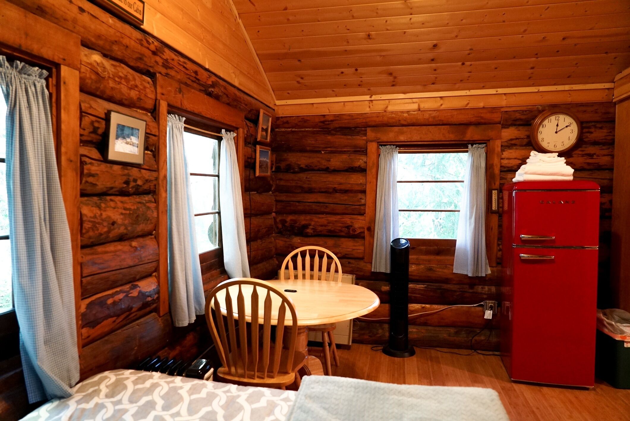 A wooden cabin interior with a wooden table, chairs, a red refrigerator, and a clock on the wall.