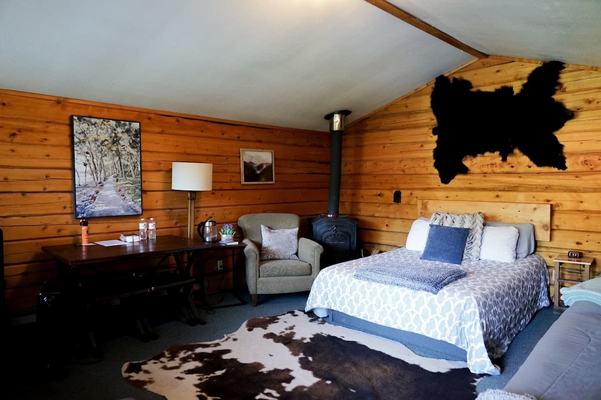 A cozy cabin room with a wooden interior, a bed with patterned bedding, a desk with a lamp, and a chair.