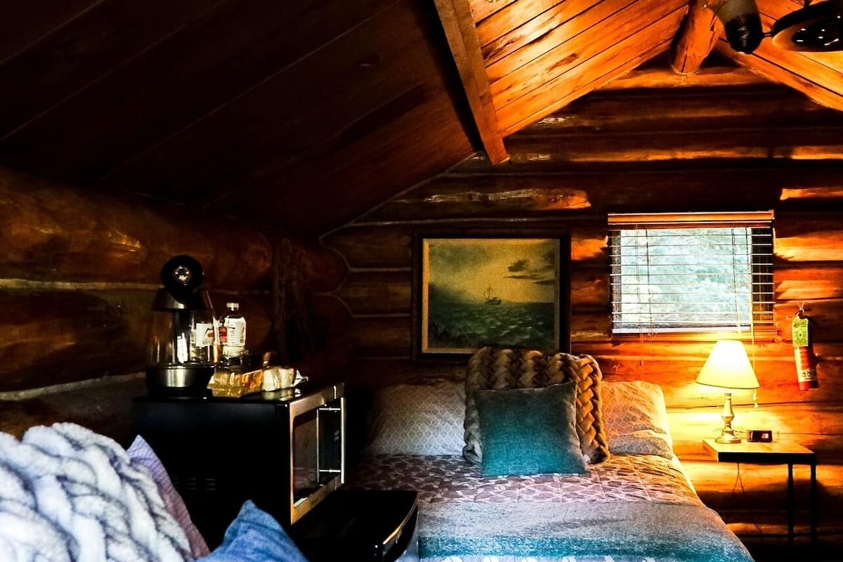 A cozy cabin room with a bed, a small table with a lamp, a painting on the wall, and a window with blinds.
