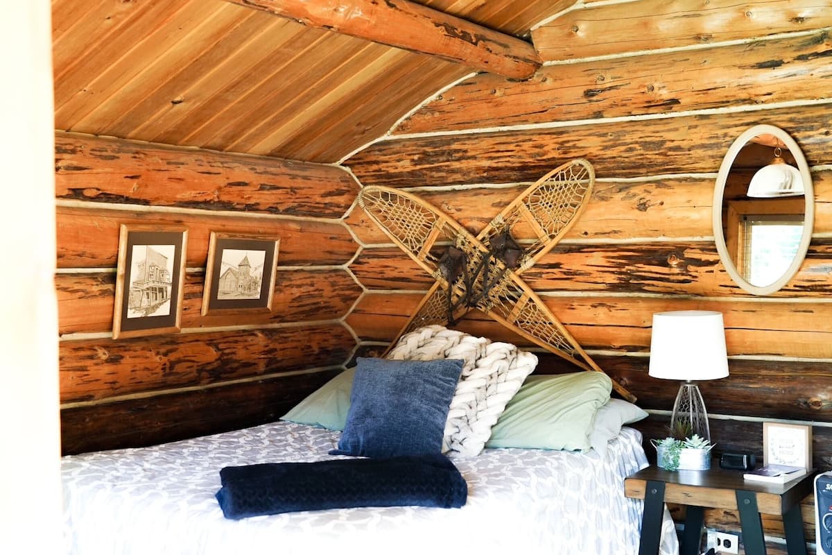 A cozy log cabin room with a bed, a woven headboard, framed pictures on the wall, a mirror, and a bedside table with a lamp and a small plant.