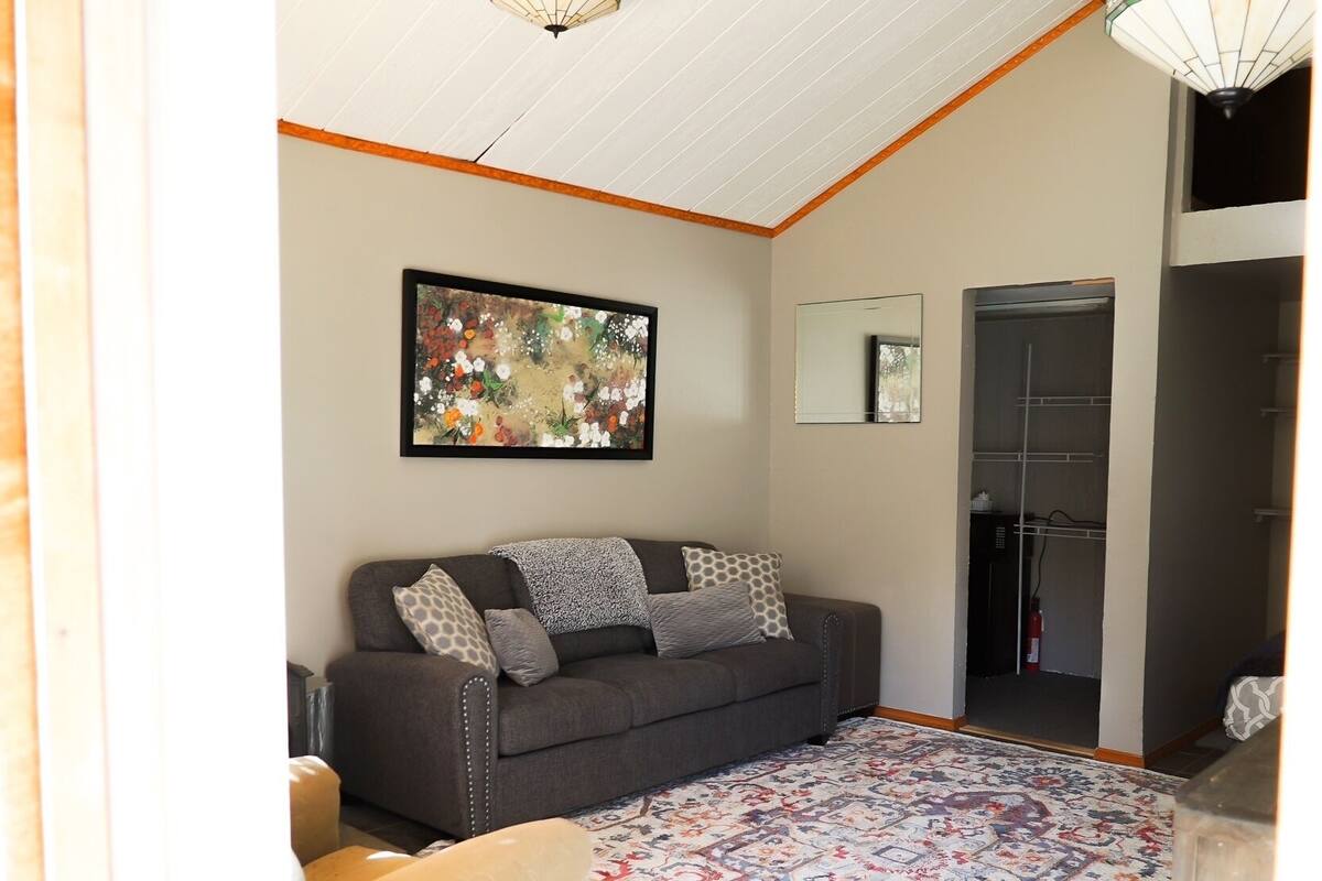 A living room with a grey sofa, patterned cushions, a large floral painting, a mirror, and a patterned rug.