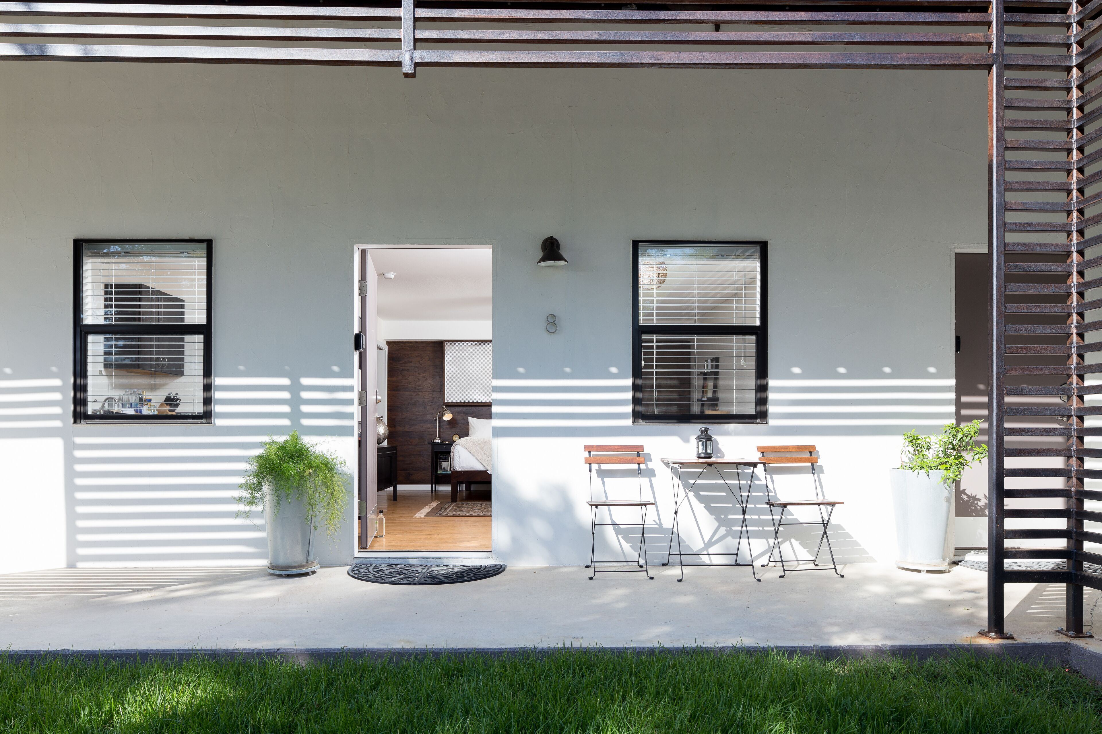 Fachada moderna blanca, dos ventanas con persianas, una puerta de acceso a una habitación, un pequeño espacio exterior con mesa y sillas, y una planta en maceta.