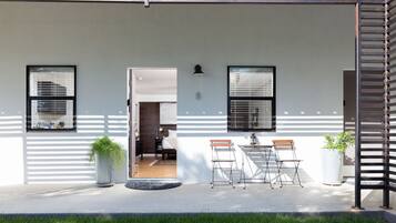 Fachada moderna blanca, dos ventanas con persianas, una puerta de acceso a una habitación, un pequeño espacio exterior con mesa y sillas, y una planta en maceta.