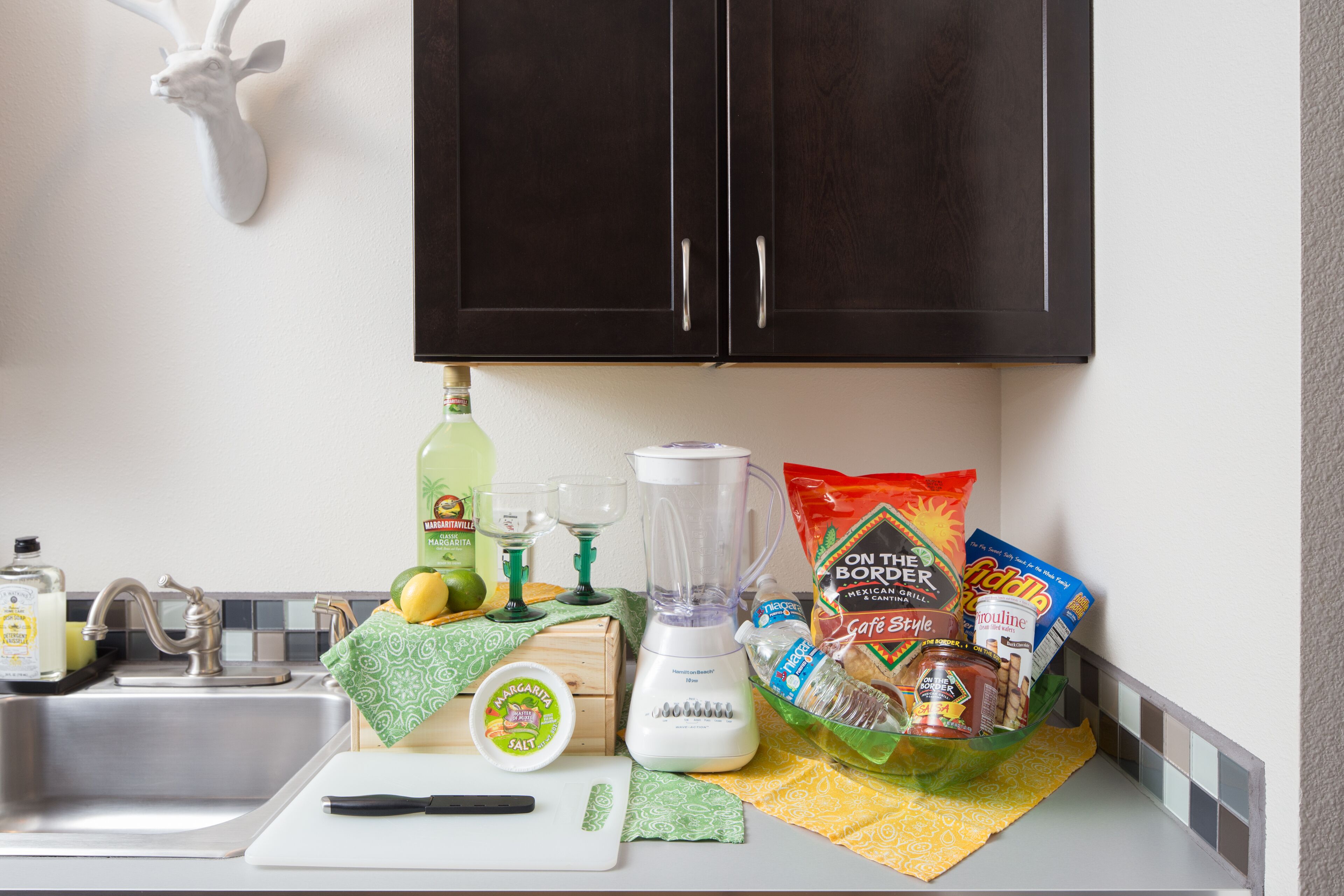 A kitchen with a sink, a cutting board, a blender, and various food items on the counter.