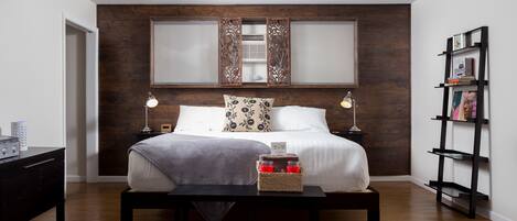 Een moderne slaapkamer met een groot bed, nachtlampen, een nachtkastje met een mand vol kaarsen, en een houten hoofdbord.