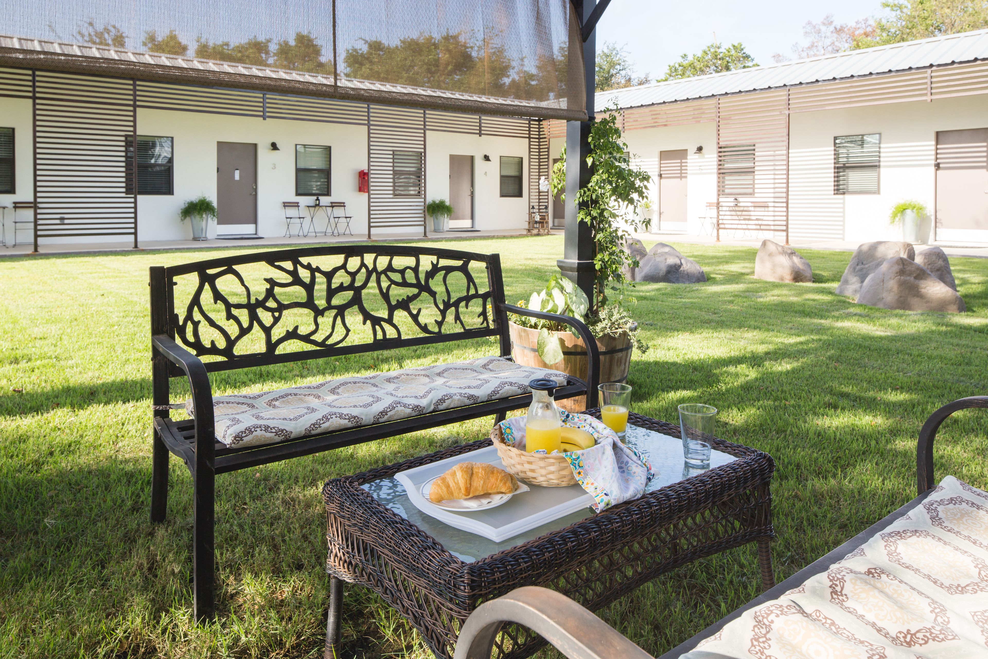 Un espace extérieur aménagé avec une table en osier, un banc et un coin petit-déjeuner comprenant des croissants, du jus de fruits et un panier de fruits.