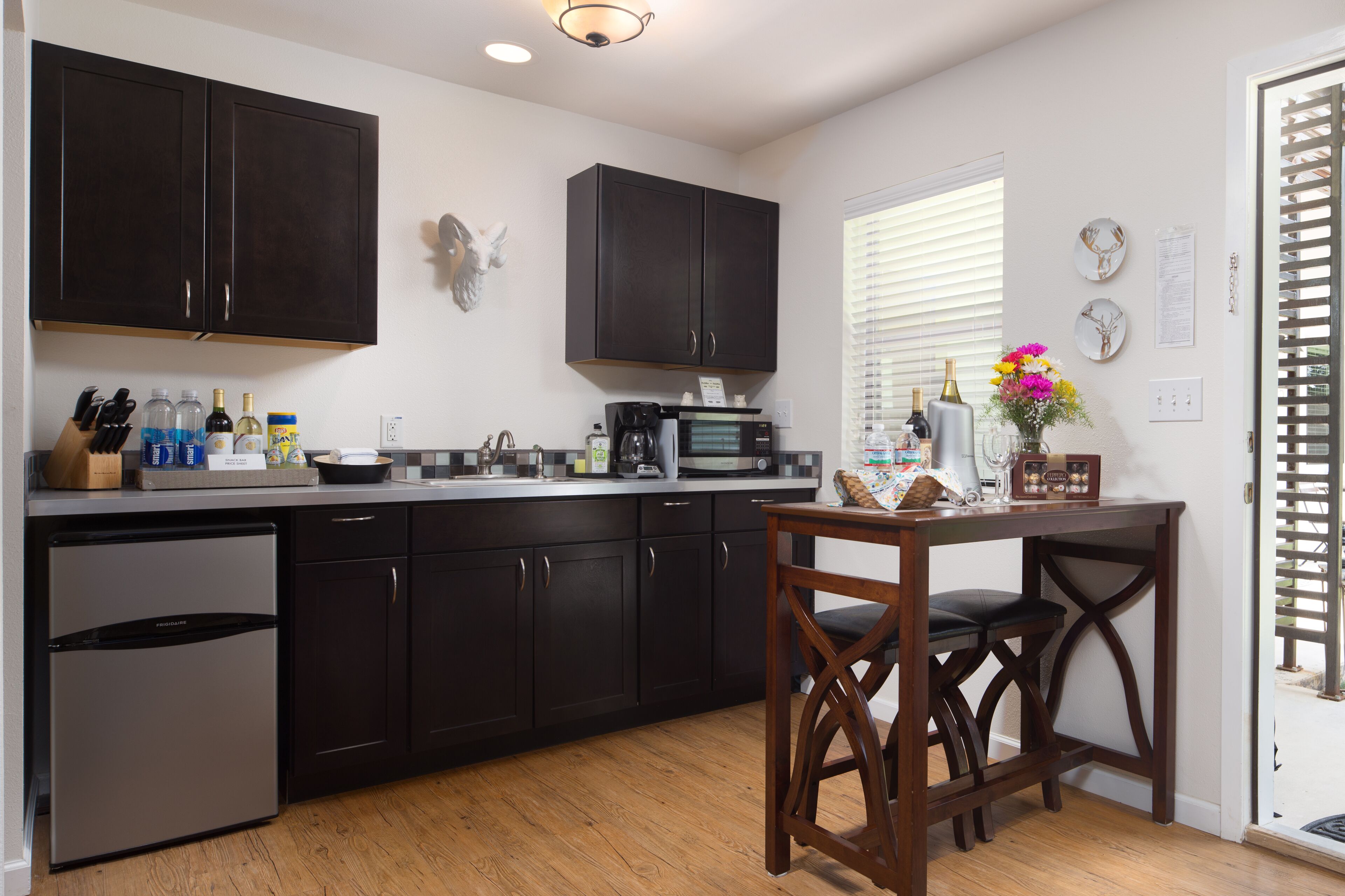 A modern kitchen with dark cabinets, a stainless steel refrigerator, a coffee maker, and a dining area with a wooden table and chairs.
