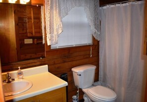 A bathroom with a wooden wall, a white sink, a toilet, and a window with a lace curtain. - Deer Haven (Blue Ridge)