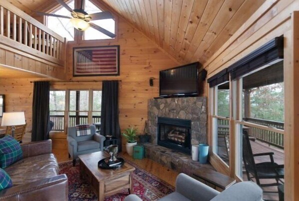 Ein gemütliches Cabin-Interieur mit Kamin aus Stein, einem Fernseher und einem Balkon mit Holzbalkon.