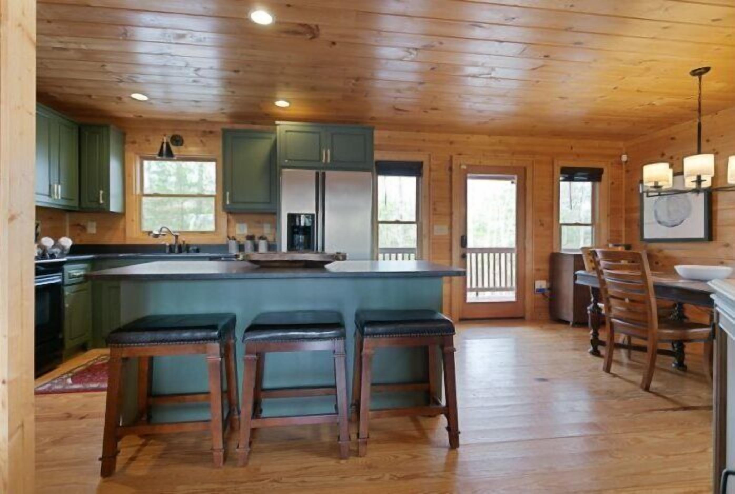A wooden cabin interior with a kitchen featuring green cabinets, a central island with stools, and a dining area with wooden chairs.