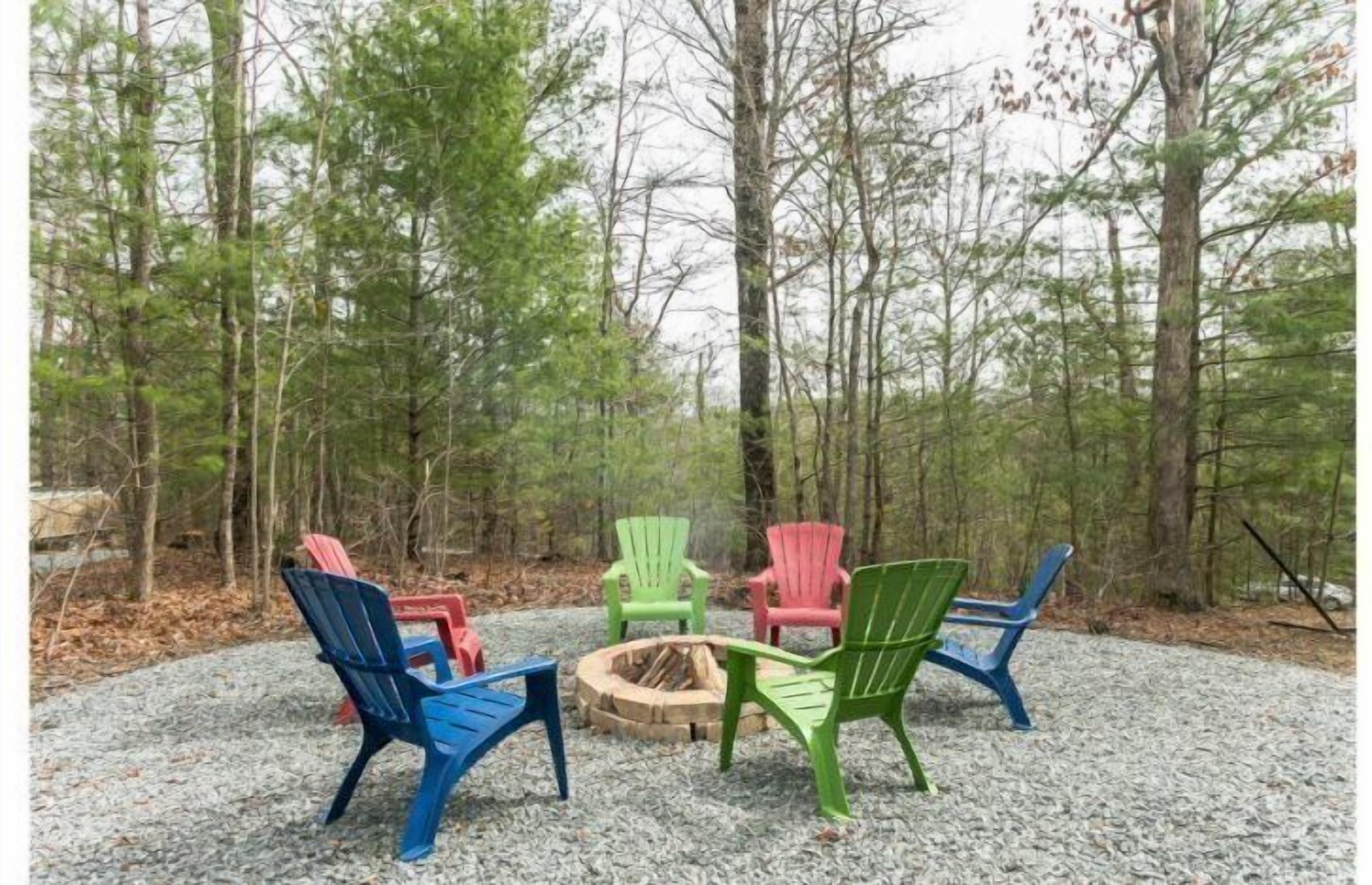 A circular seating area with Adirondack chairs around a central fire pit, set on gravel in a forested area.