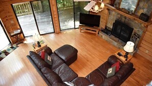 Un salon chaleureux avec un canapé d’angle, un téléviseur à écran plat, une cheminée et une grande fenêtre donnant sur une terrasse.