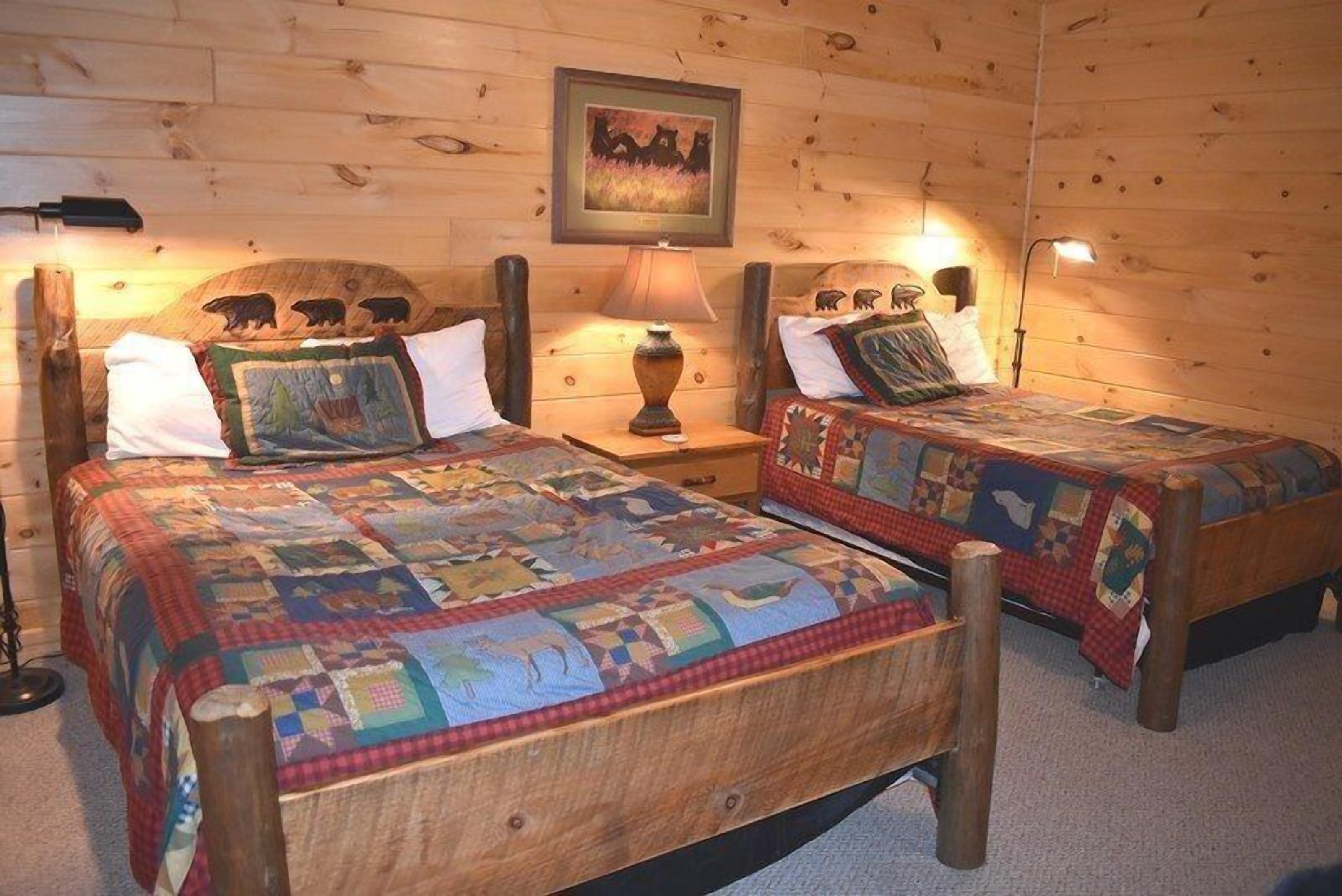 A cozy cabin-style room with two wooden beds, each with a quilted bedspread and matching headboards. There is a wooden wall with a bear carving and a framed picture above the beds.