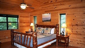 Une chambre dans une cabane en bois, avec un lit, deux tables de chevet, un ventilateur de plafond et une fenêtre avec des volets.