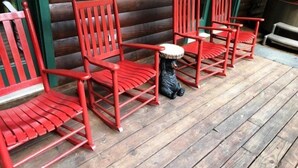 En rad röda trästolar med en liten svart björnfigur placerad mellan dem, ställda på ett trädäck framför en timmerstuga.