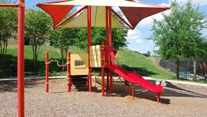 A playground with a red slide and a yellow climbing structure, surrounded by trees and a grassy hill in the background.
