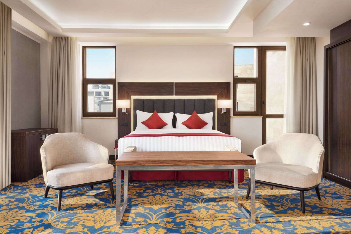 Une chambre d’hôtel avec un grand lit, deux fauteuils, une table en bois et une moquette à motifs.