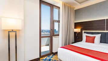 A hotel room with a large bed, a view of the city through a window, a floor lamp, and a carpet with a blue and yellow pattern.